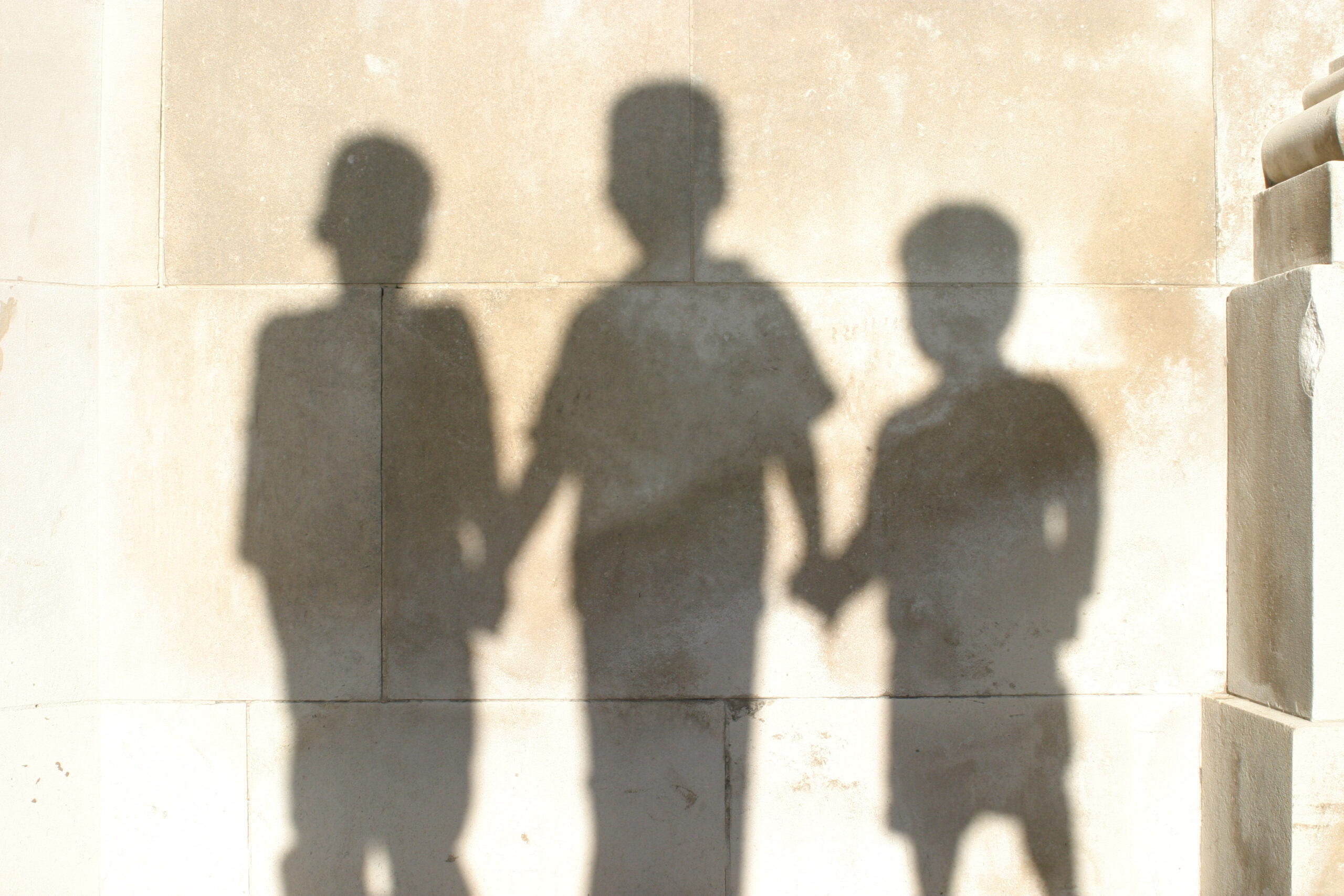 Shadows of three children holding hands, standing near a wall during the day.